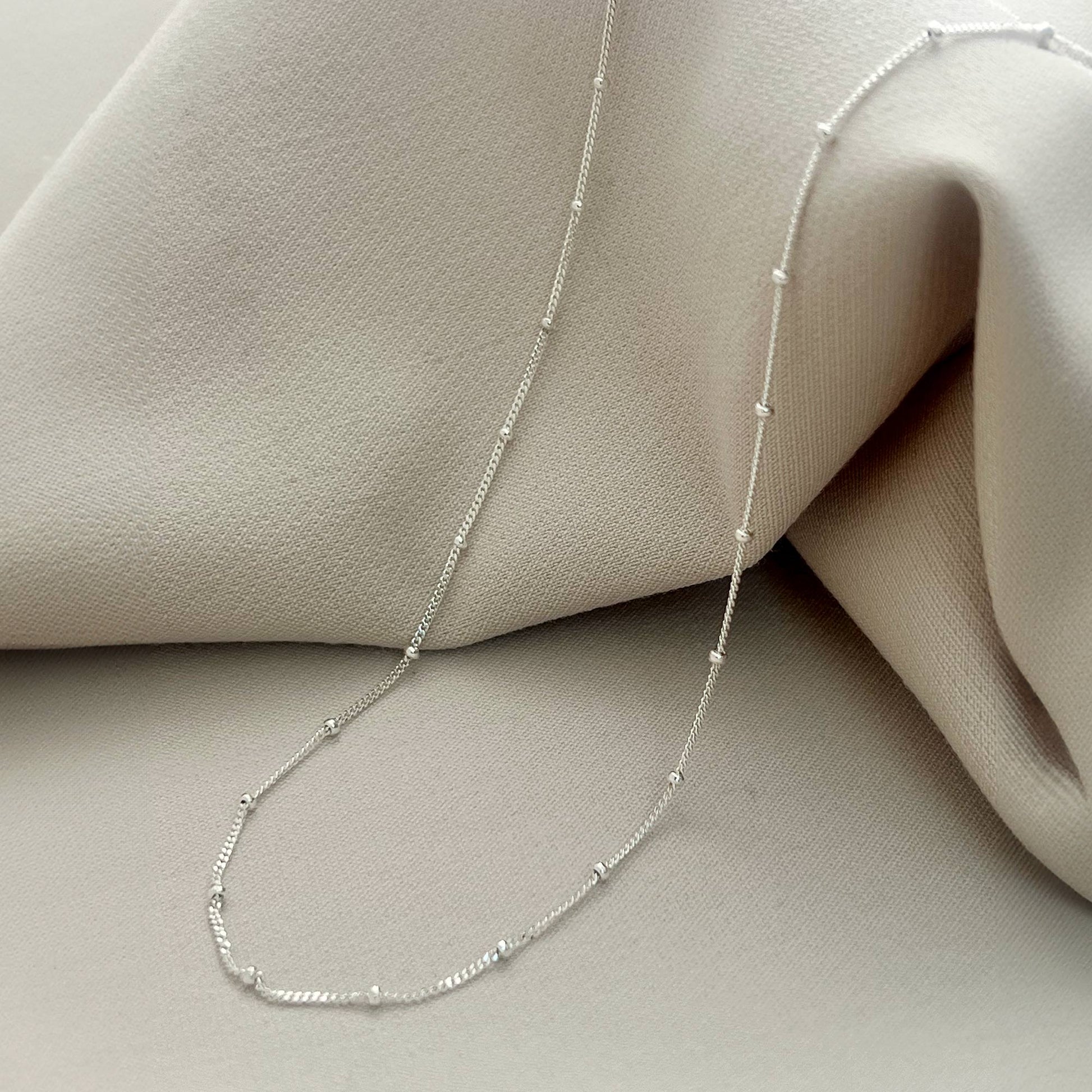 Close-up of Dainty Bead Chain Necklace Silver against a soft beige fabric backdrop.