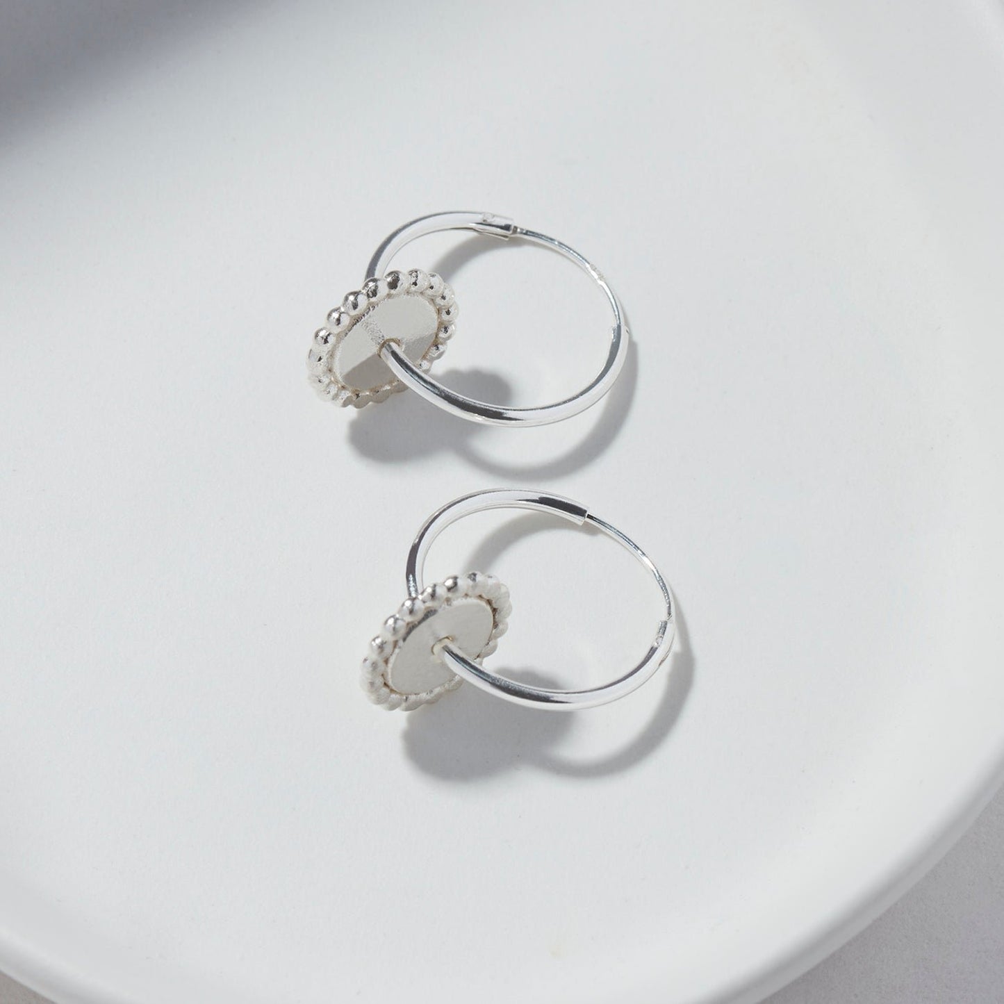 Close-up of silver hoop earrings with a dotted, removable circular disc detail on a white plate.