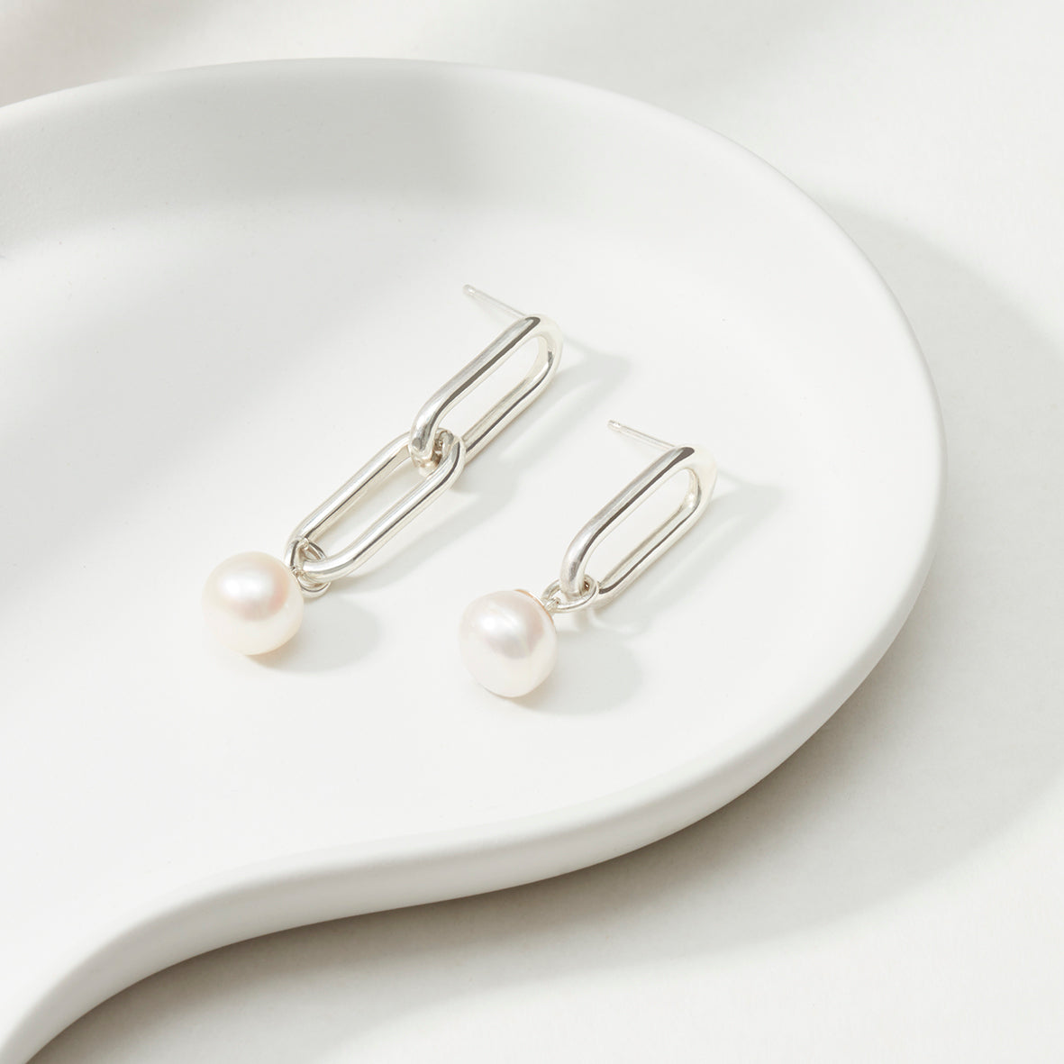 Close-up of mismatched silver link earrings featuring a freshwater pearl drop, resting on a white dish.