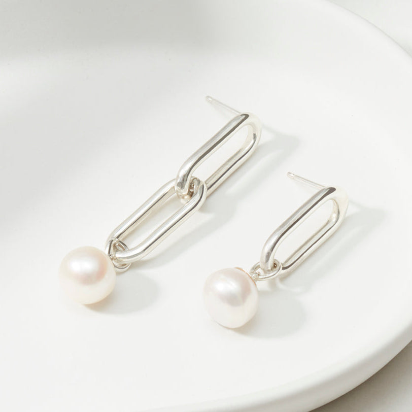 Close-up of mismatched silver link earrings featuring a freshwater pearl drop, resting on a white dish.